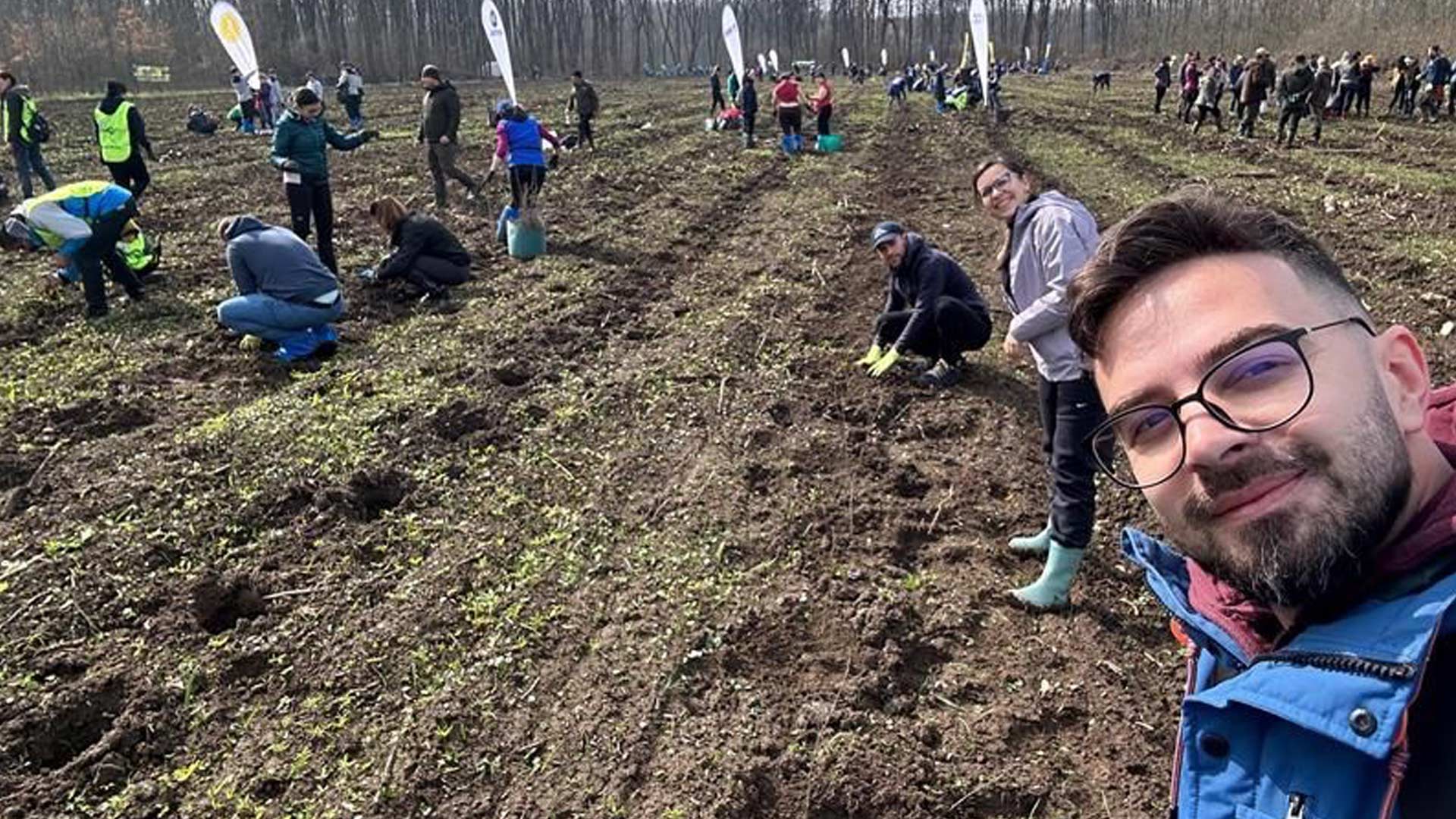Group of people planting trees in a field, wearing casual outdoor clothing. A person in glasses takes a smiling selfie in the foreground, conveying a cheerful and collaborative atmosphere.