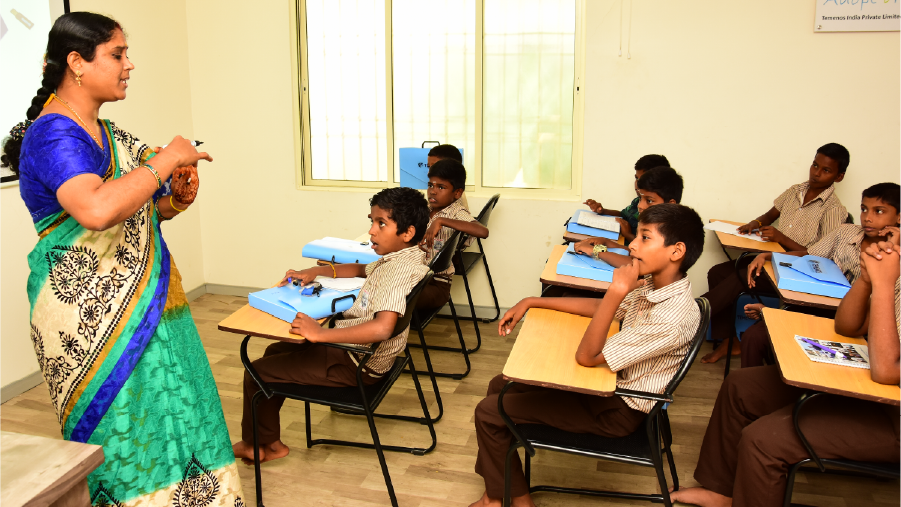 A teacher in a colorful sari engages with attentive students in a classroom. Boys in striped uniforms sit at desks with blue folders, creating a focused learning atmosphere.