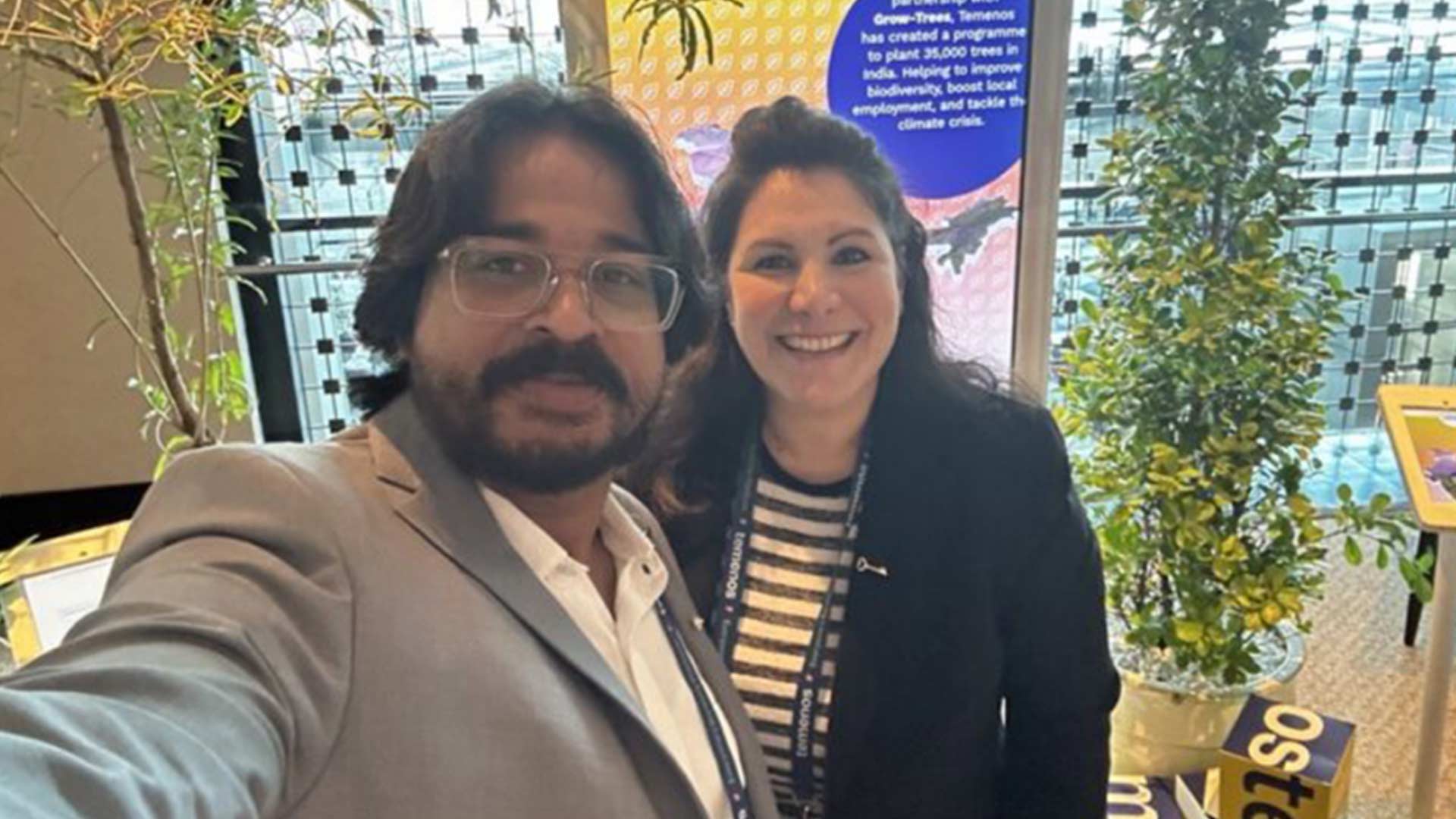 Two people smiling in a modern indoor setting with greenery. They're in front of a colorful poster about tree planting for biodiversity and climate action.