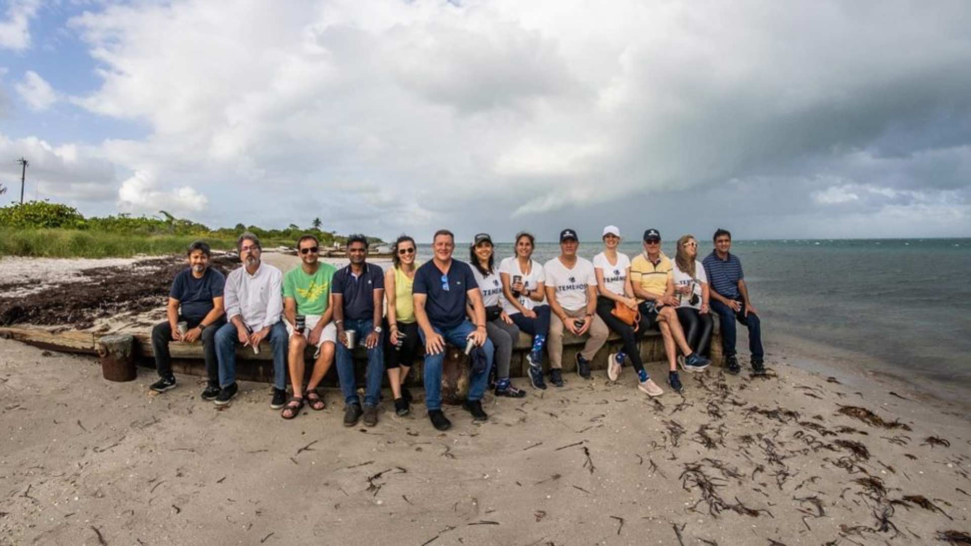 A group of 13 people sit on a log at a beach, smiling under a partly cloudy sky. The ocean is on their right, and grass is visible on the left.