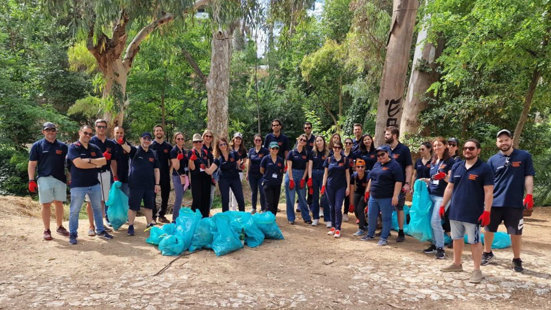 A group of people in matching dark blue shirts and red gloves stand together in a park. They are surrounded by trees and carry blue garbage bags, suggesting a community cleanup effort. The mood is positive and collaborative.