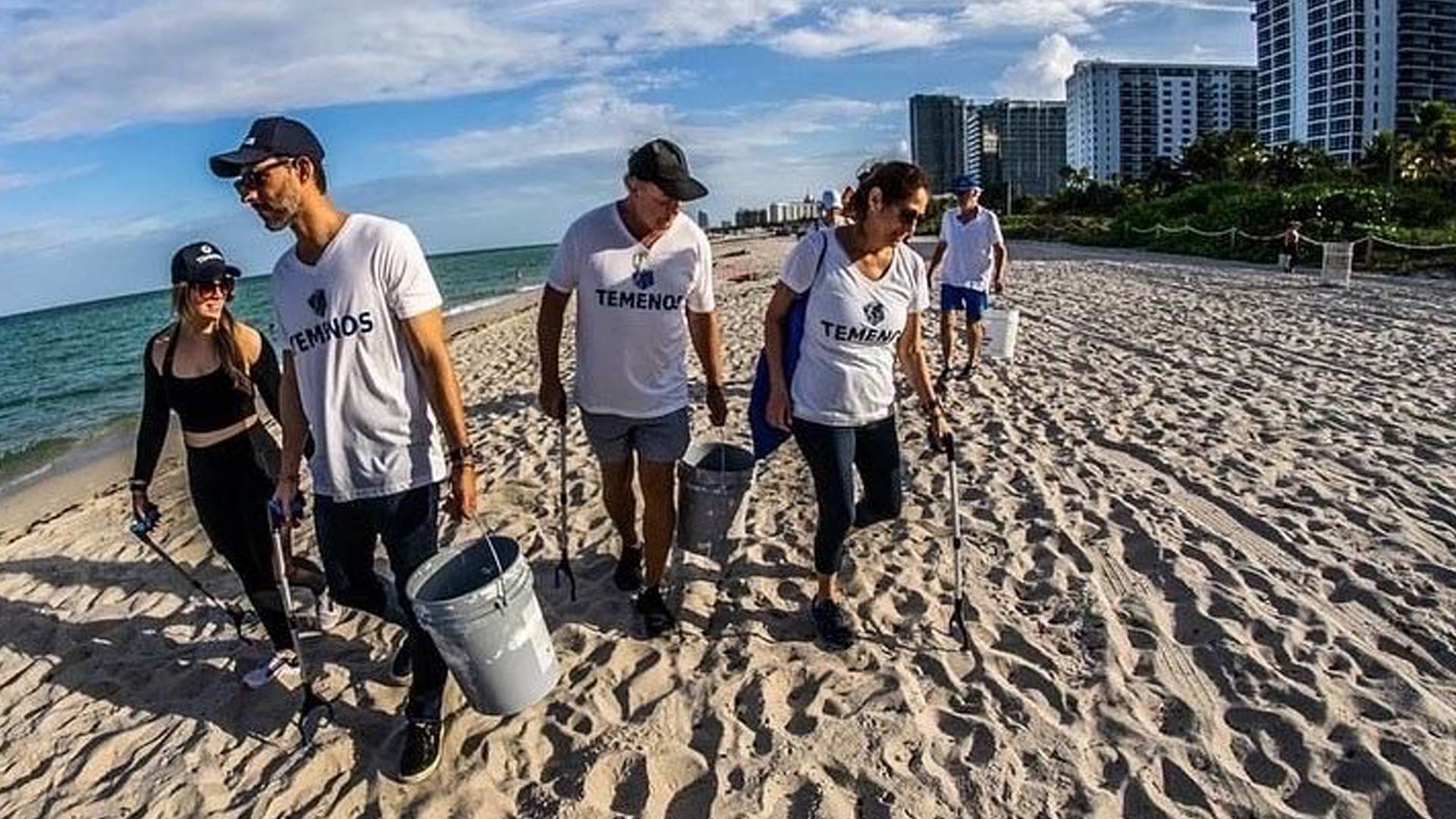 A group of people, wearing casual clothes and "TEMENOS" shirts, joyfully clean a sunny beach, holding buckets and litter pickers. Tall buildings are visible in the background under a partly cloudy sky.
