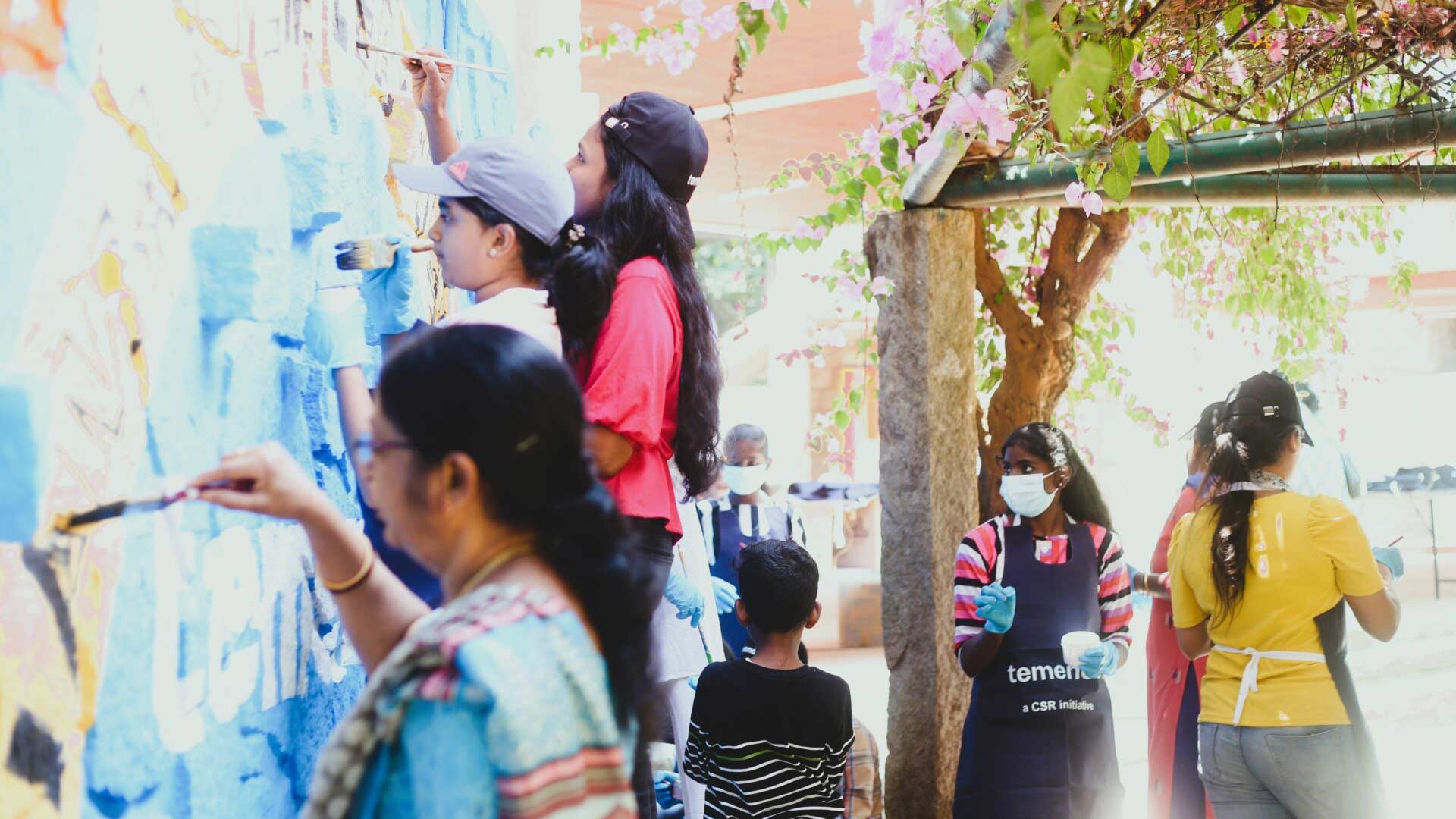 A group of people, including children, paint a mural on a wall outdoors. They are focused and wearing casual clothes and aprons. Flowers and trees surround them, creating a lively, collaborative atmosphere.