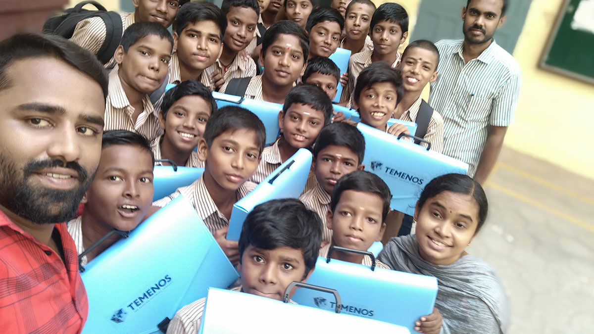 A joyful group of schoolchildren and two teachers pose with blue folders labeled "Temenos." They are smiling, creating a cheerful and lively atmosphere.