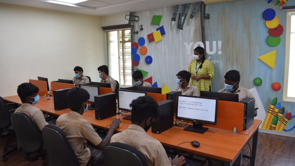 Students wearing masks sit at computers in a vibrant classroom with geometric art on the wall. An instructor stands nearby overseeing the session.