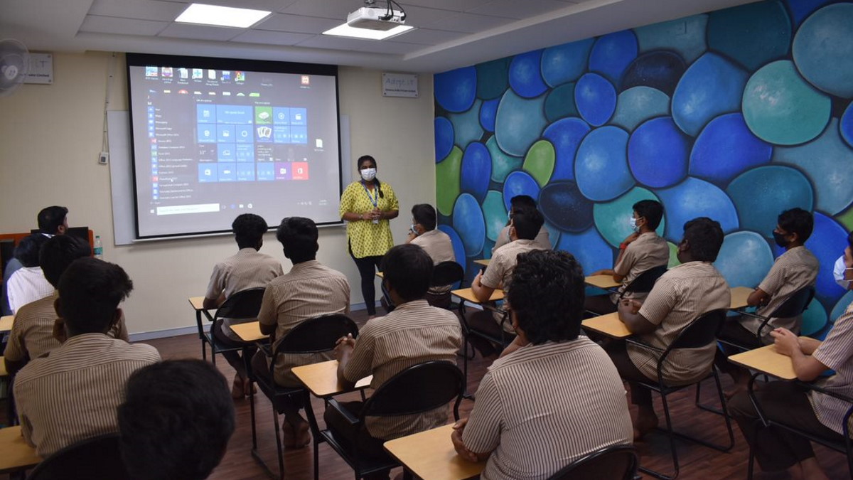 A classroom setting with a teacher wearing a yellow shirt and mask standing near a projector screen displaying a computer desktop. Students in striped uniforms sit at desks, facing the teacher. The blue and green patterned wall creates a vibrant backdrop. The atmosphere is attentive and focused.