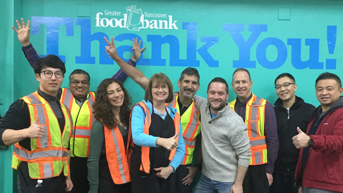 A diverse group of nine smiling people wearing reflective vests stands in front of a "Thank You!" sign at Greater Vancouver Food Bank, radiating positivity.