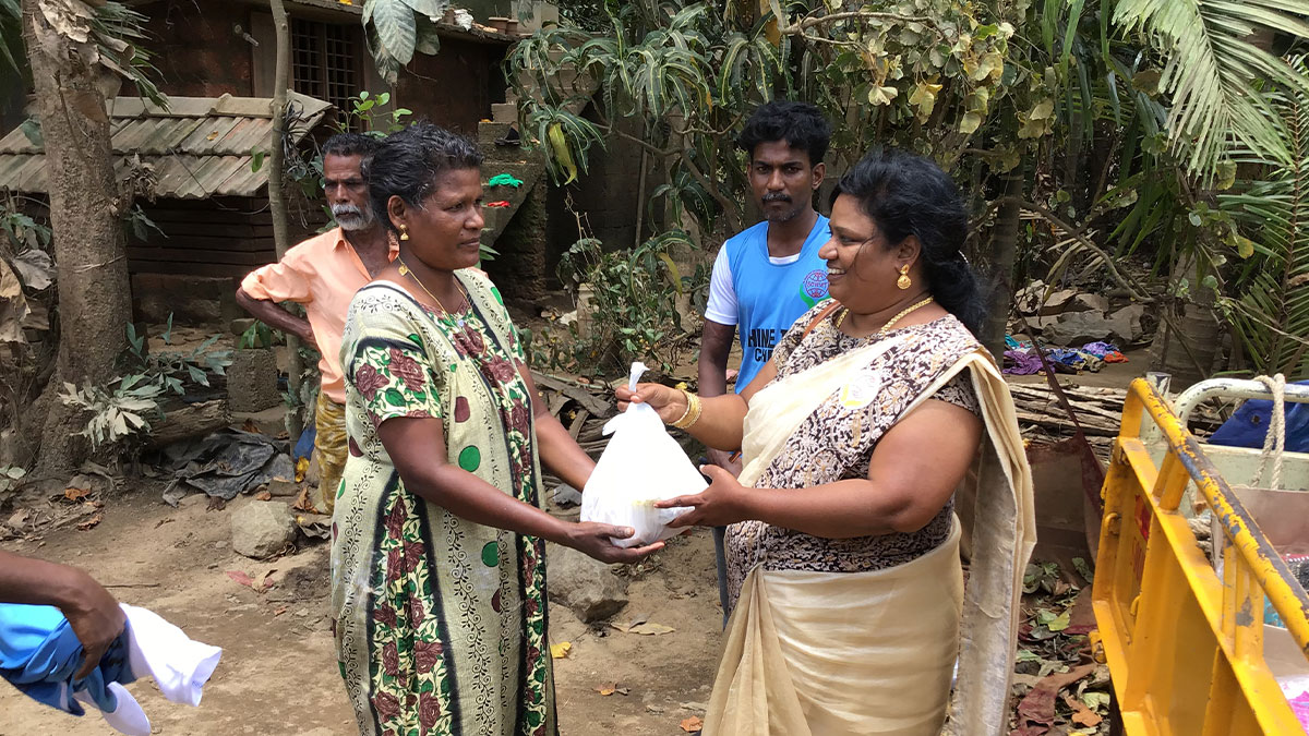A woman in traditional attire smiles while handing a white bag to another woman in a patterned dress. Bystanders and a lush, rural setting are in the background.