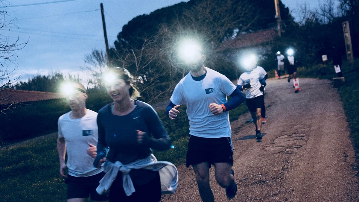 Runners with headlamps jog on a dimly lit rural road, evoking a sense of camaraderie and determination in the early morning or dusk ambiance.