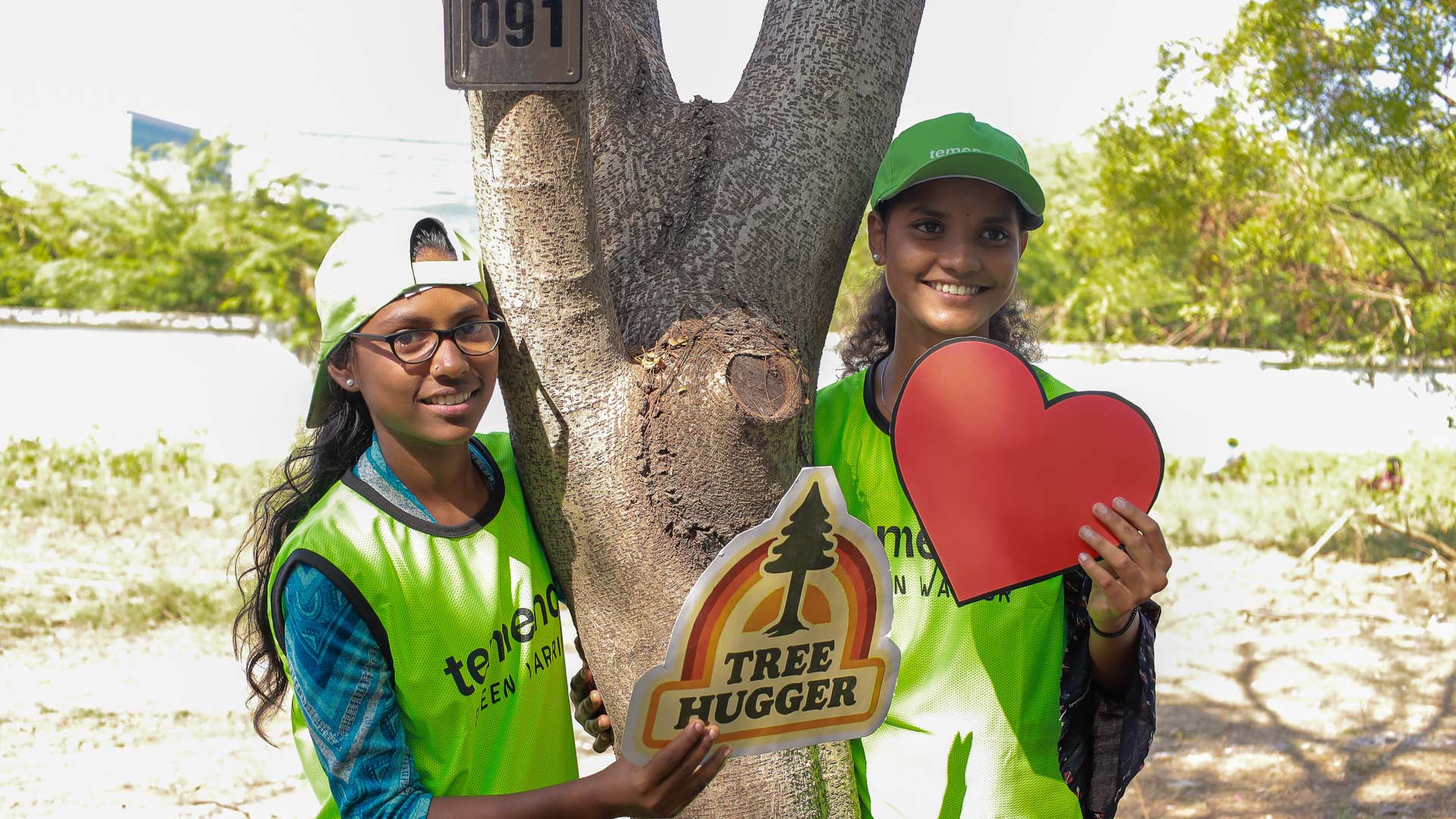 Two smiling people in green vests and caps stand by a tree, holding signs: one reads "Tree Hugger," the other is a red heart. Bright, sunny day.