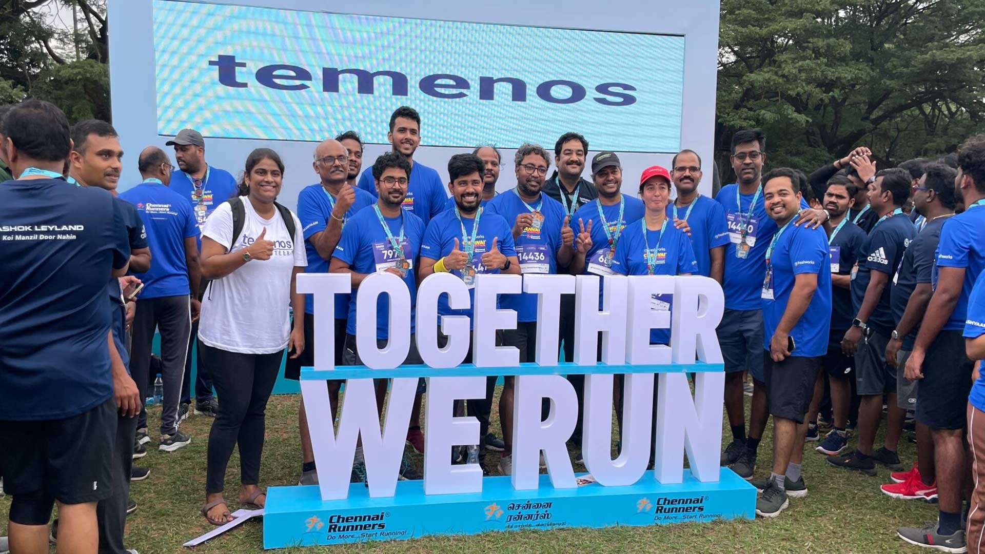 A large group of people, diverse in appearance and attire, pose together indoors against a green wall with a logo. The mood is friendly and inclusive.