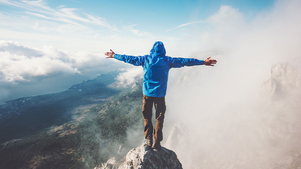 Person in a blue jacket stands on a mountain peak with arms outstretched, surrounded by fog and clouds, conveying freedom and triumph.