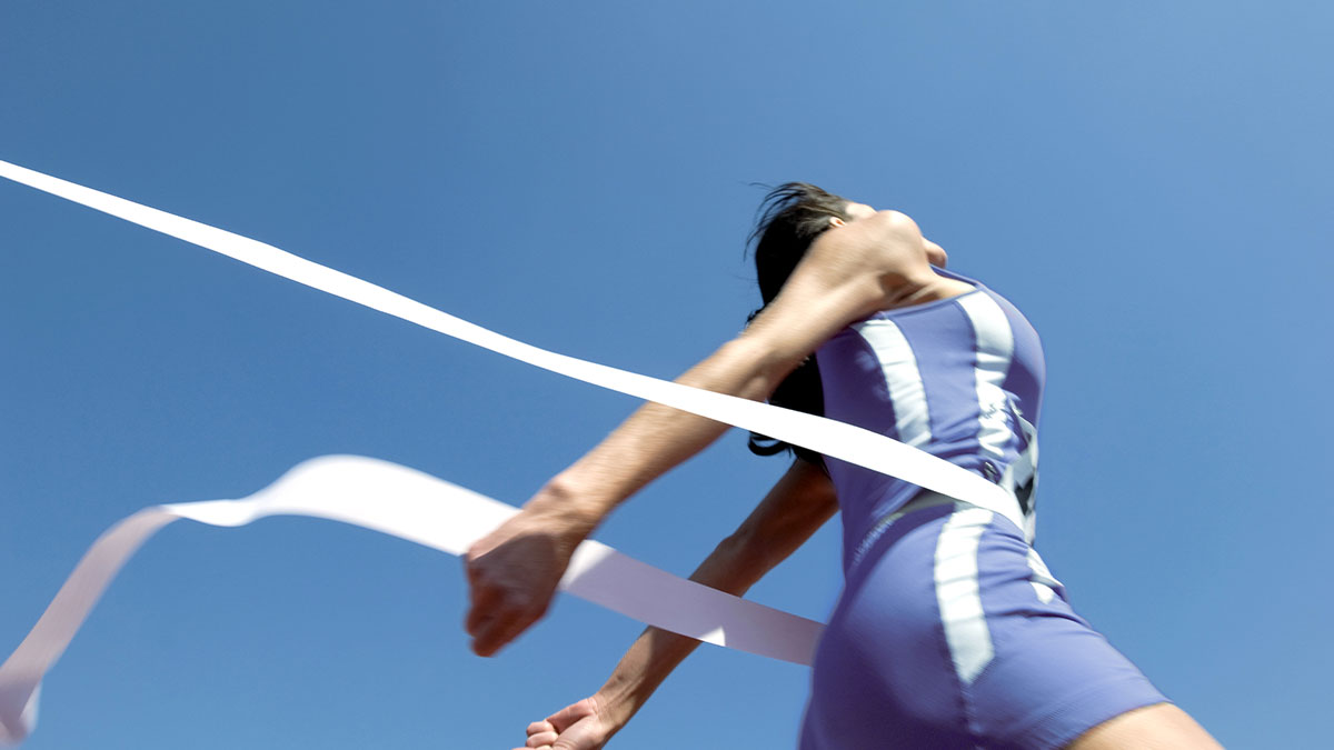 A runner in a blue and white suit crosses the finish line with arms wide, capturing a moment of triumph against a clear blue sky.