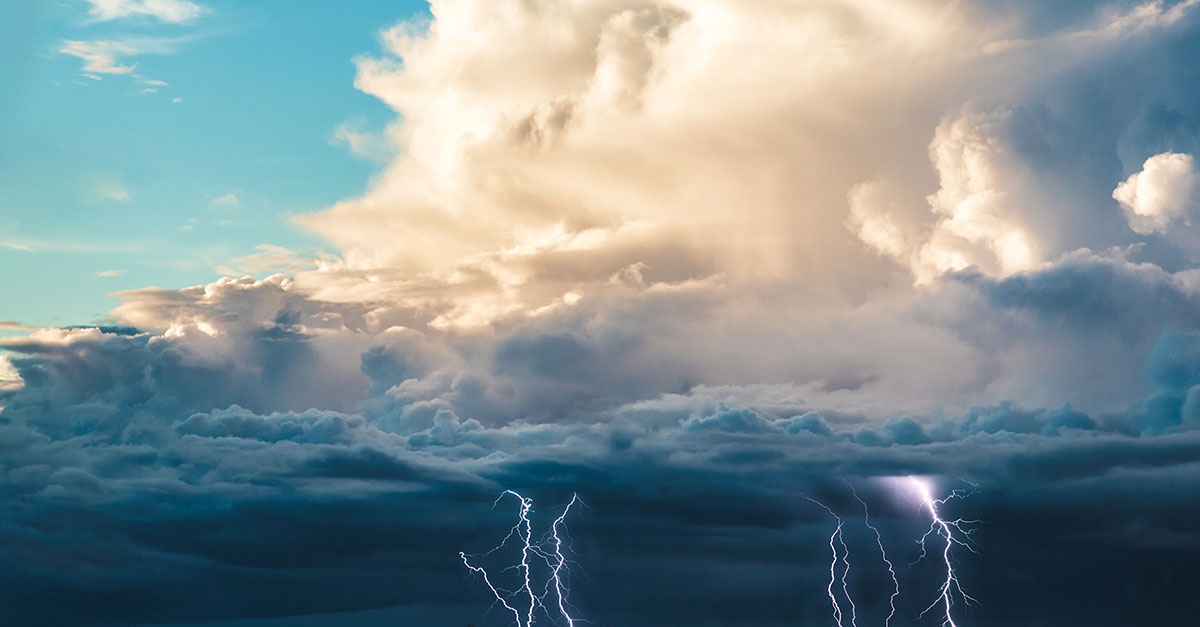 Dramatic sky with large, billowing clouds illuminated by sunlight against a blue backdrop. Bright lightning strikes emerge below, conveying intensity.