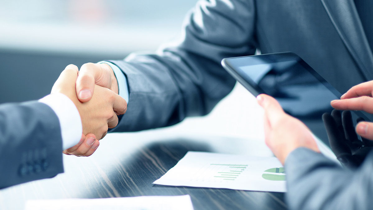 Two professionals in suits shake hands over a table with a tablet and documents showing graphs, conveying a successful business agreement.