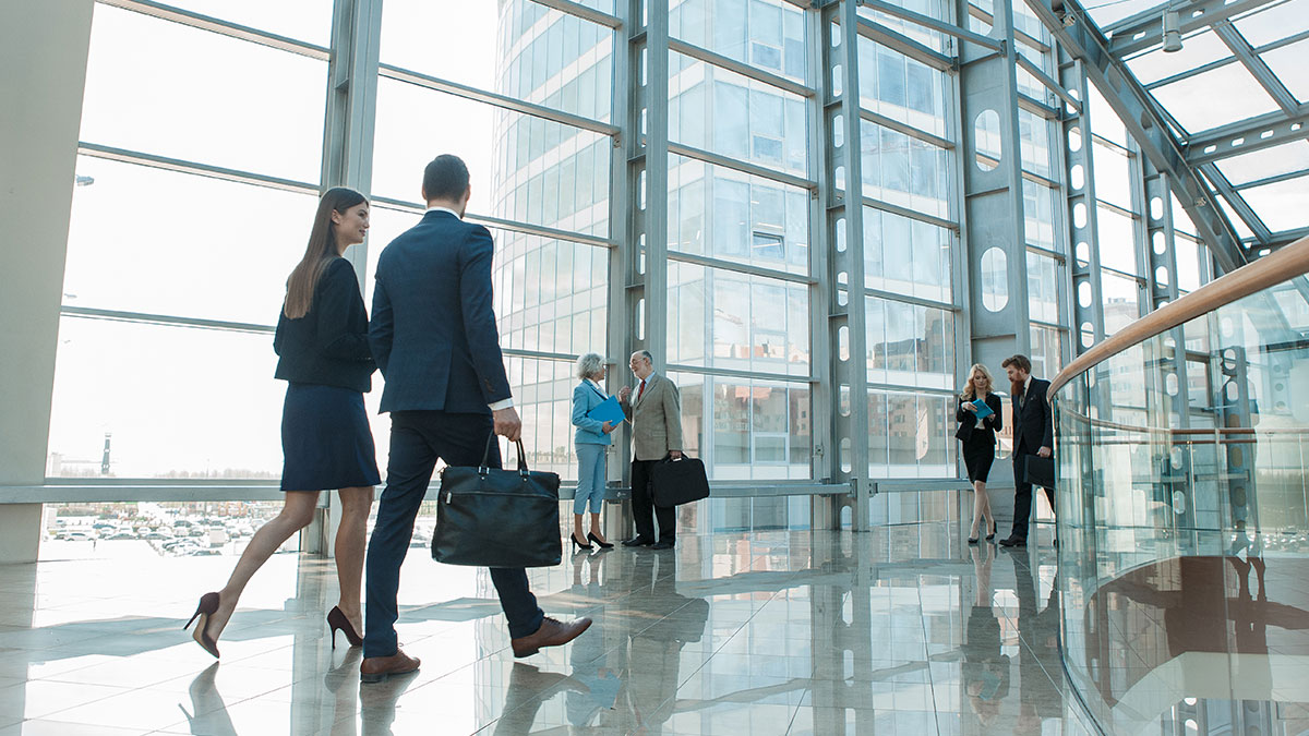 Business professionals in formal attire walk and converse in a spacious, sunlit office atrium with large windows and shiny floors, conveying a busy, collaborative atmosphere.