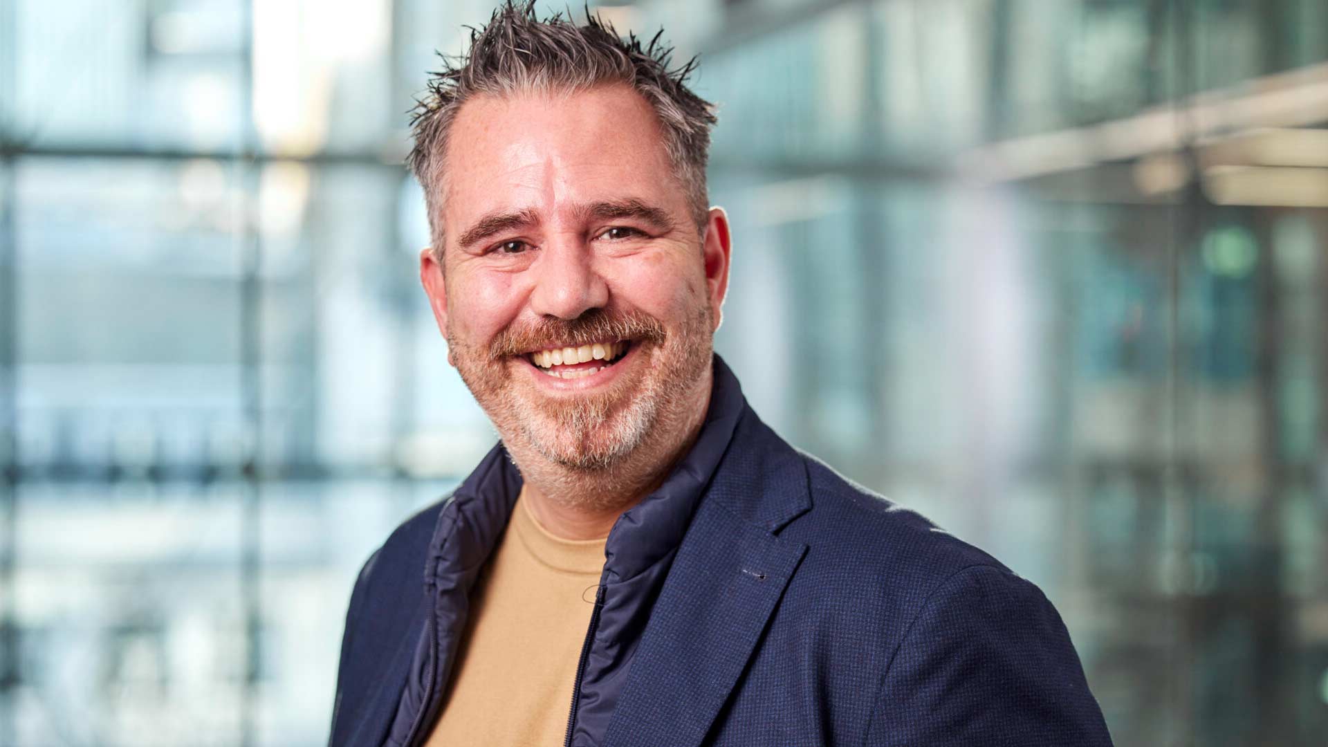 Smiling man with short, spiked hair in a blue blazer and beige shirt. Bright, modern office background with large windows, conveying a cheerful tone.
