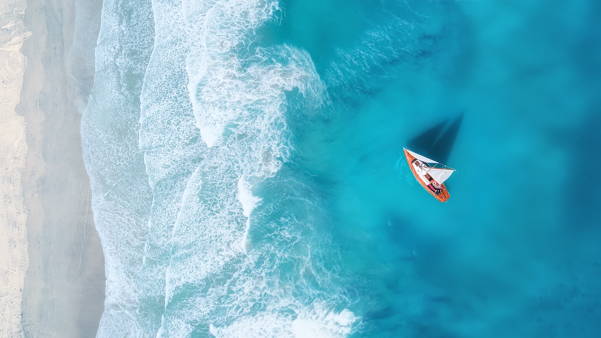 Aerial view of a small sailboat on vibrant blue water near a sandy beach. White waves crash gently onto shore, creating a peaceful, serene scene.