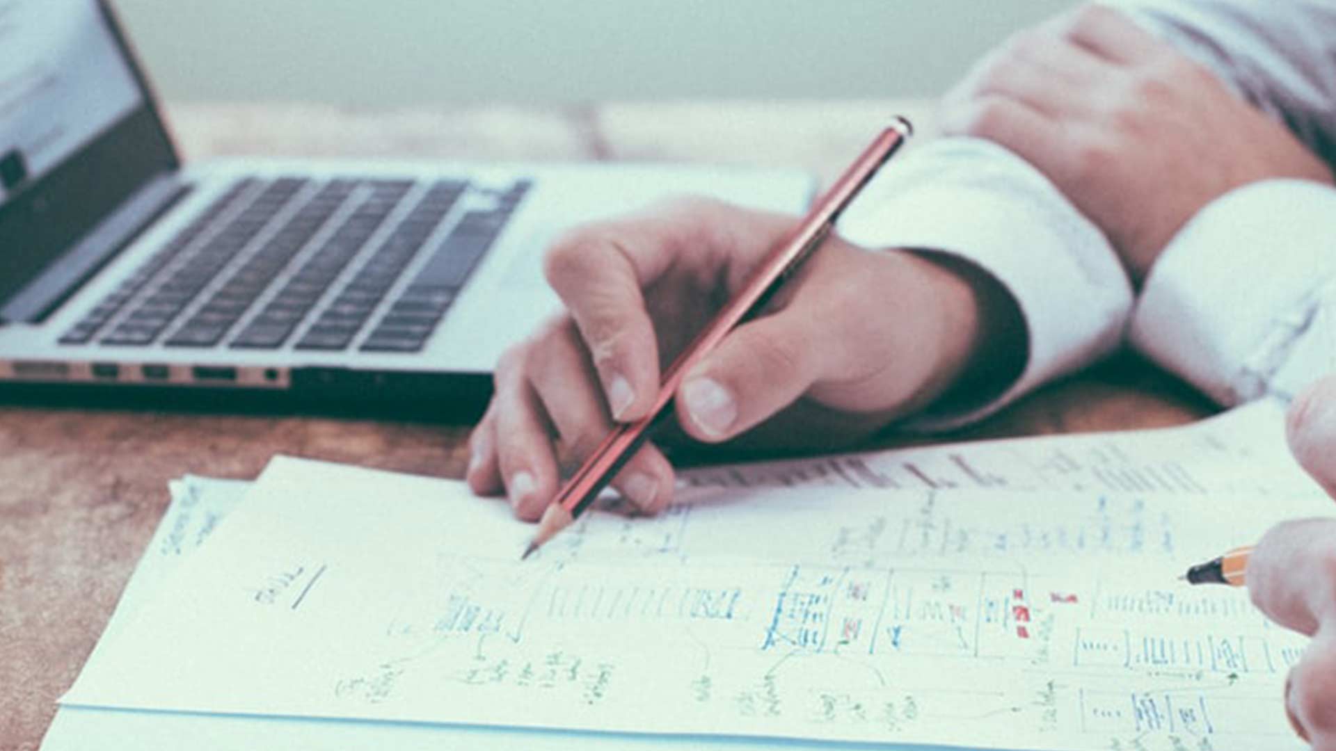 Hands writing on papers with diagrams beside a laptop. The scene conveys focus and planning, with a collaborative and professional tone.