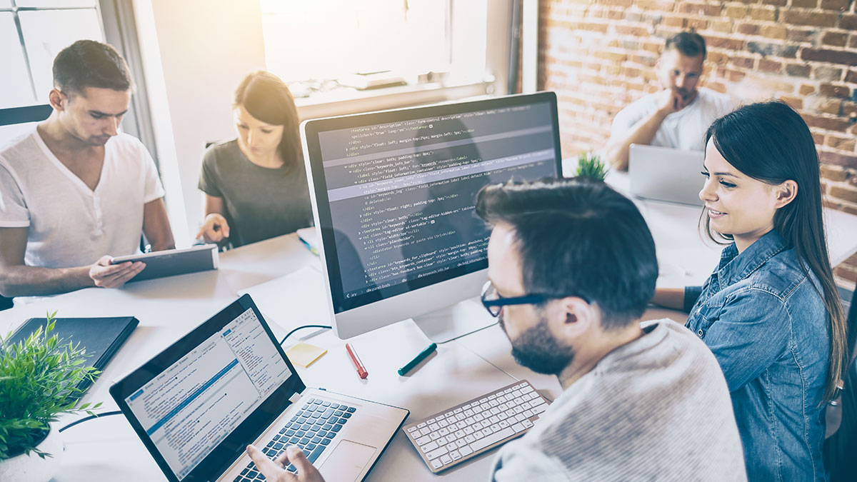 A group of five people work collaboratively in an office with computers displaying code. The room is lit by natural sunlight, creating a productive atmosphere.