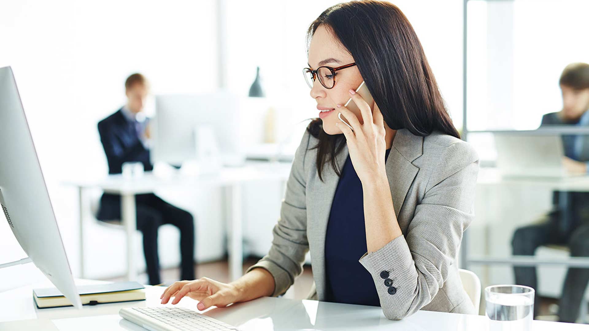 A woman in a gray blazer, wearing glasses, is on the phone and typing on a keyboard in a bright office. Two colleagues work at their desks in the background.