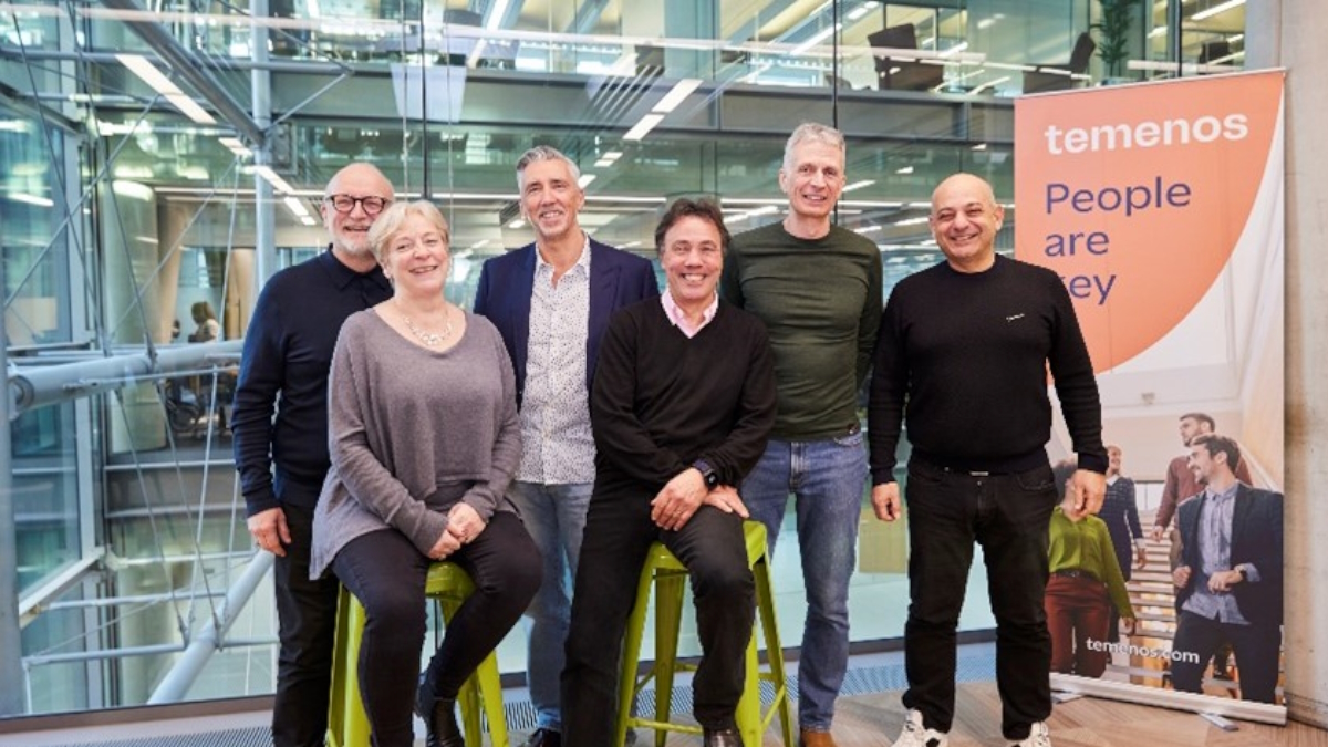 Six smiling people, casually dressed, pose in a modern office with glass walls. A Temenos banner reads, "People are key." Positive and professional atmosphere.