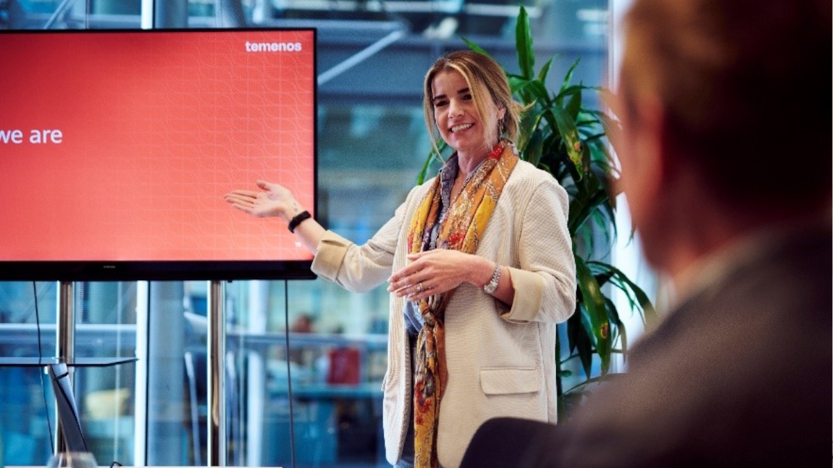 A woman in a white blazer and scarf presents enthusiastically beside a red screen displaying "temenos." A blurred audience member listens intently.