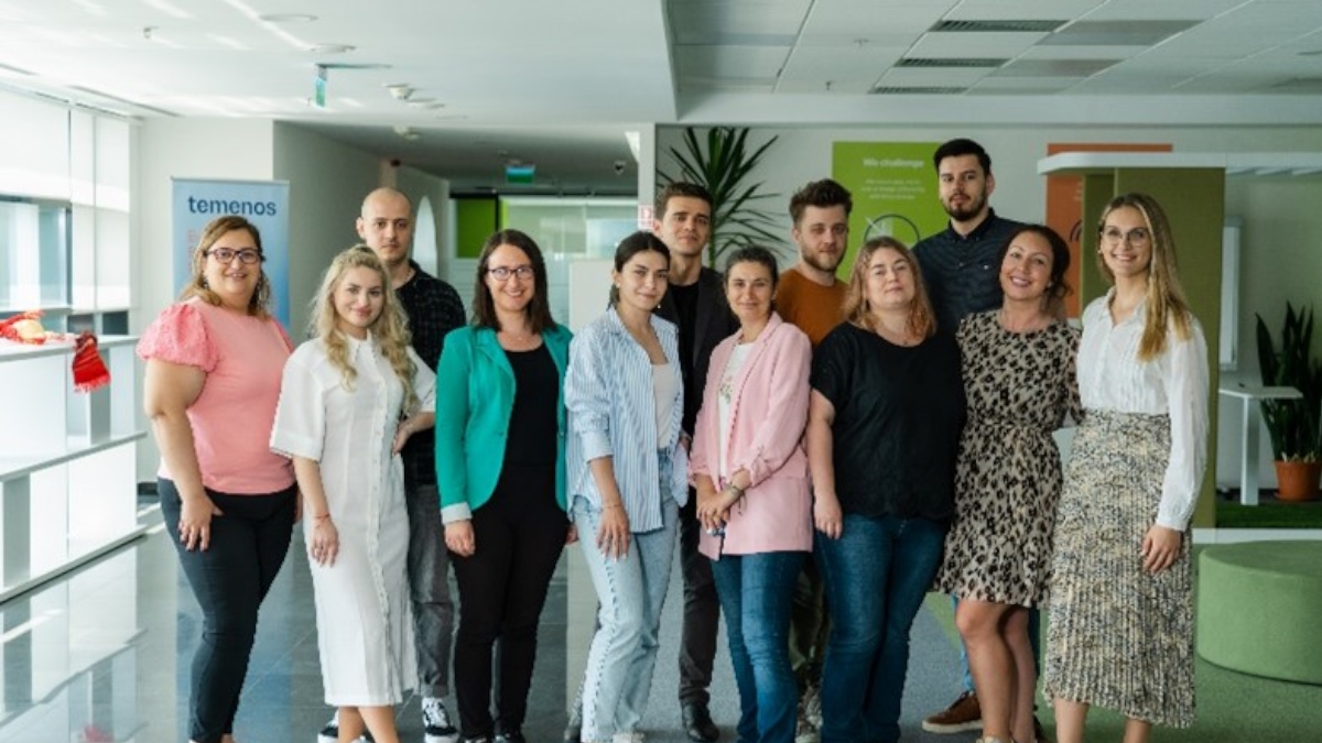 A group of 11 diverse people standing together in an office hallway, smiling warmly. Bright, modern space with plants and motivational posters. Friendly atmosphere.