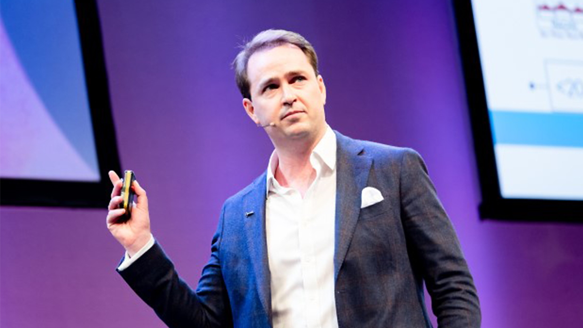 Man in a suit holds a phone on stage, addressing an audience. He appears confident and focused. Background shows a purple-lit screen.