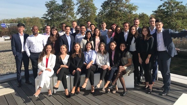 A diverse group of young professionals in business attire smile for a photo outdoors on a sunny day with trees and a blue sky in the background.