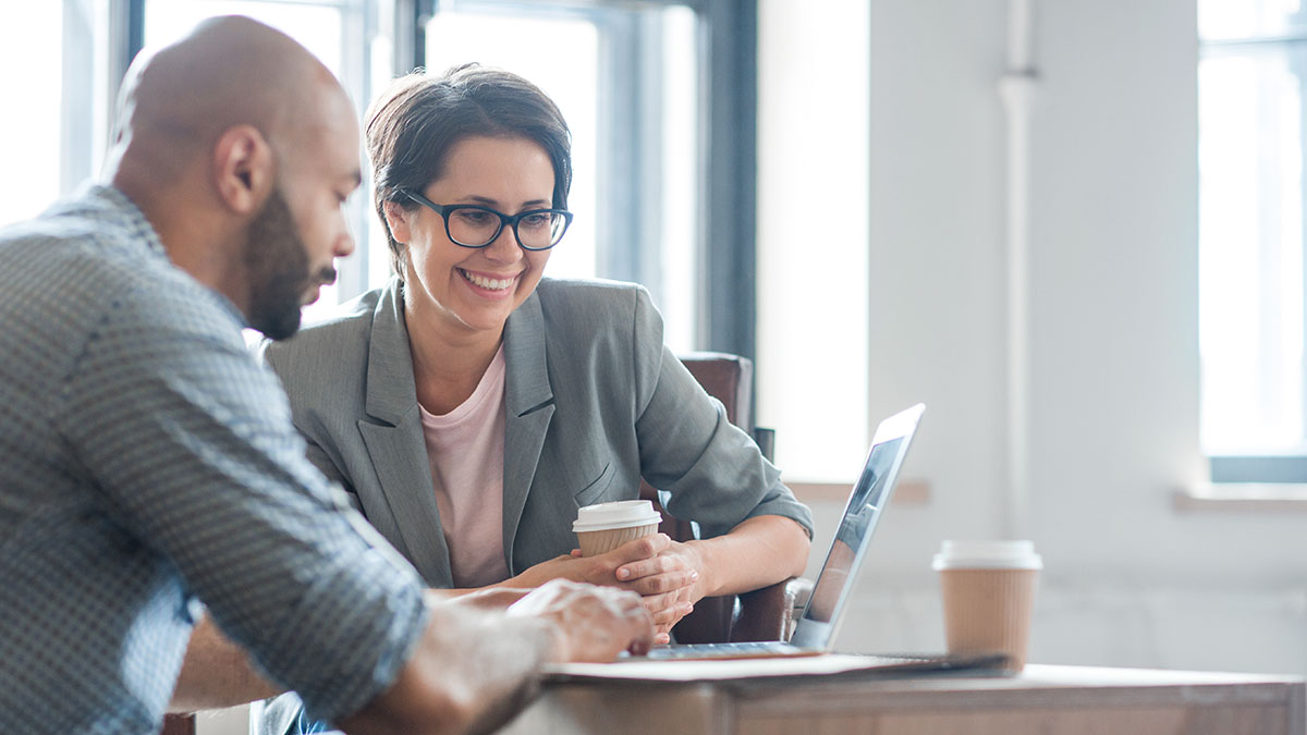 Two colleagues sit at a table, smiling and discussing work over coffee. A laptop is open in front of them, creating a collaborative atmosphere.