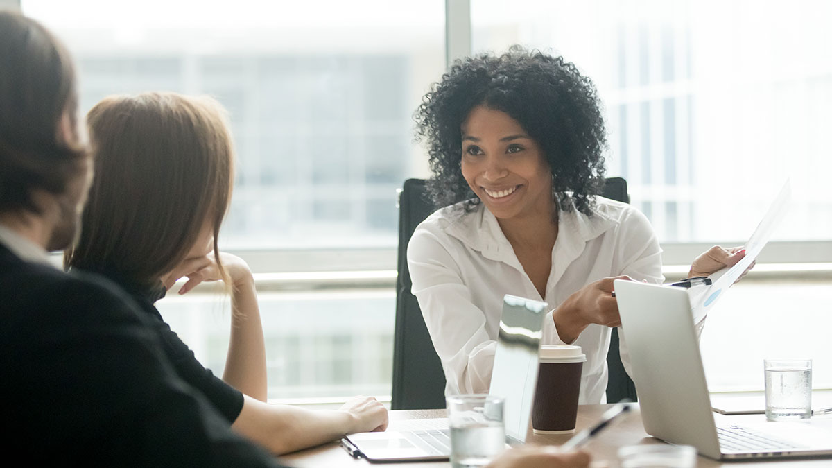 A woman in a white shirt, smiling, presents a document during a meeting. Two colleagues listen attentively around a table with laptops and coffee cups.