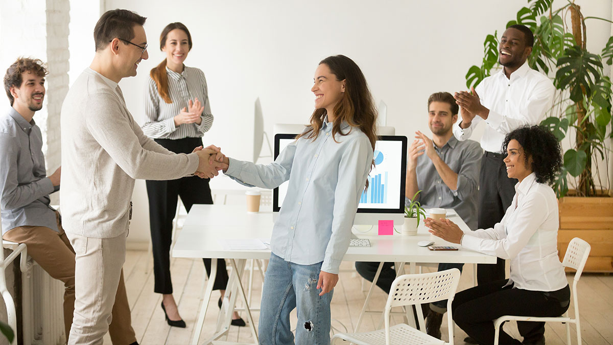 A diverse group in an office applauds as a man and woman shake hands, symbolizing teamwork. A computer displays charts. The mood is joyful and collaborative.