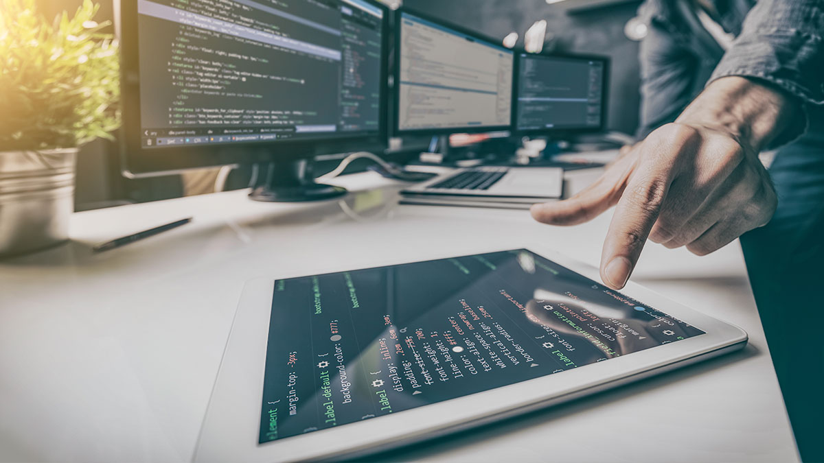 A person points at a tablet displaying colorful code, surrounded by multiple computer screens showing code. The scene conveys a focused tech workspace.