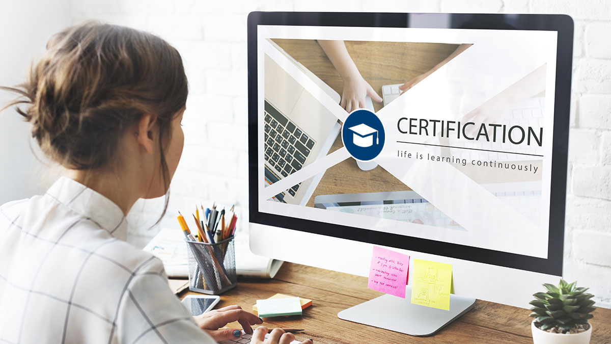 Woman at desk looks at computer screen displaying "Certification" with a graduation cap icon. Desk has colorful sticky notes and stationery, conveying a focused, educational atmosphere.