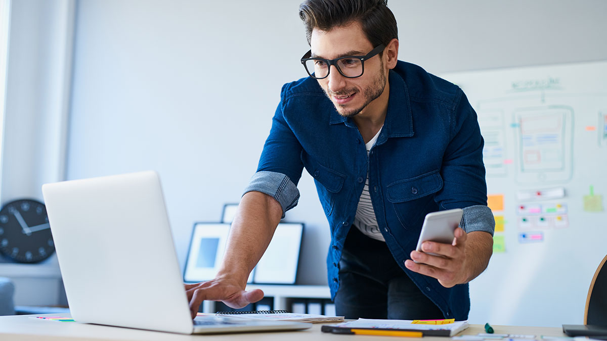 A man with glasses works on a laptop, holding a smartphone, in a modern office. A whiteboard with diagrams is in the background, conveying focus.