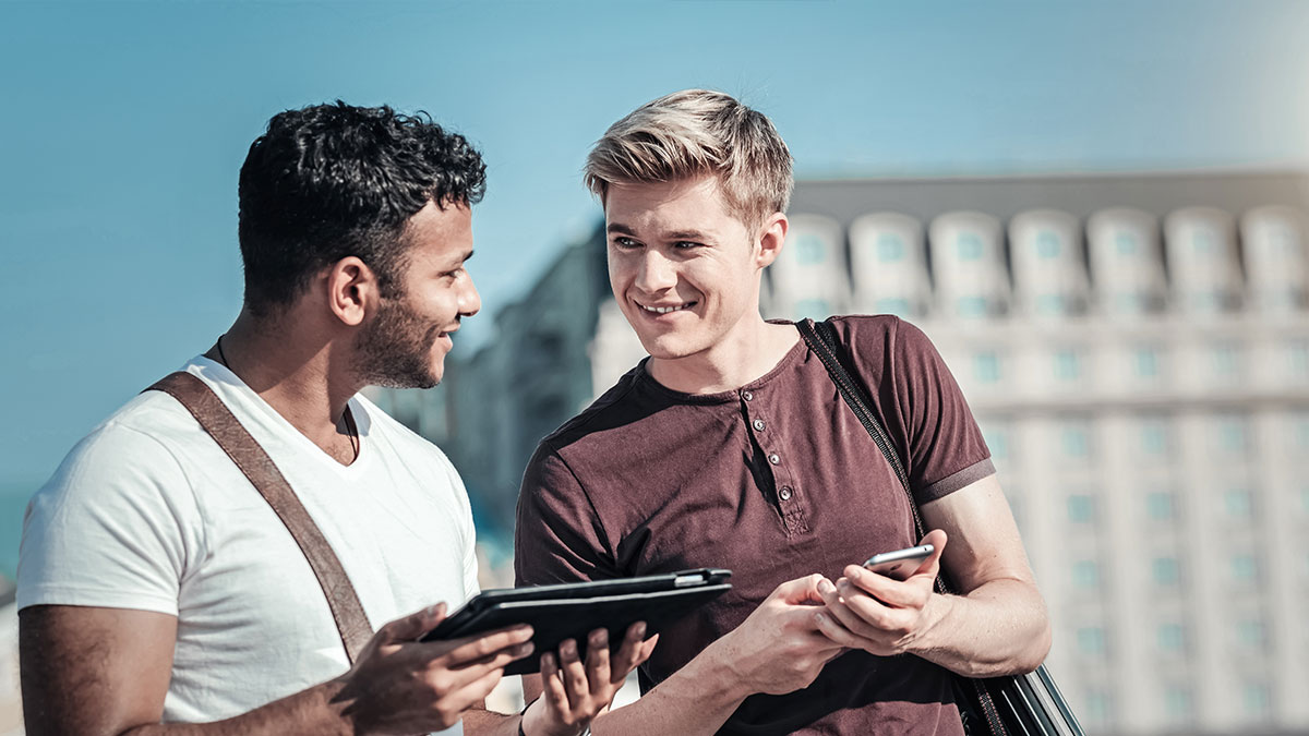 Two men stand outdoors, smiling and talking. One holds a tablet, the other a phone. The background shows a blurred building under a bright sky.