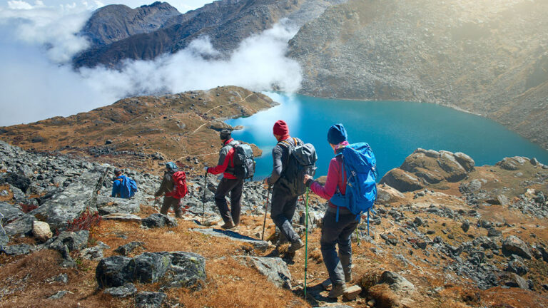 Four hikers with backpacks and trekking poles descend a rocky mountain path, overlooking a serene blue lake surrounded by mist and rugged peaks.