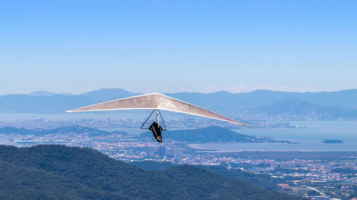 Hang glider soars over a scenic coastline with distant cityscape and mountains under a clear blue sky.
