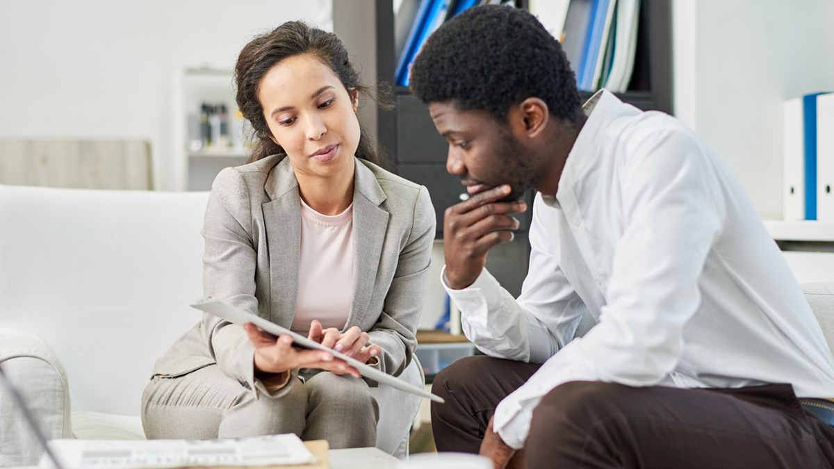 A woman in a gray suit shows a man in a white shirt something on a tablet. They appear focused, sitting on a sofa in a professional setting.