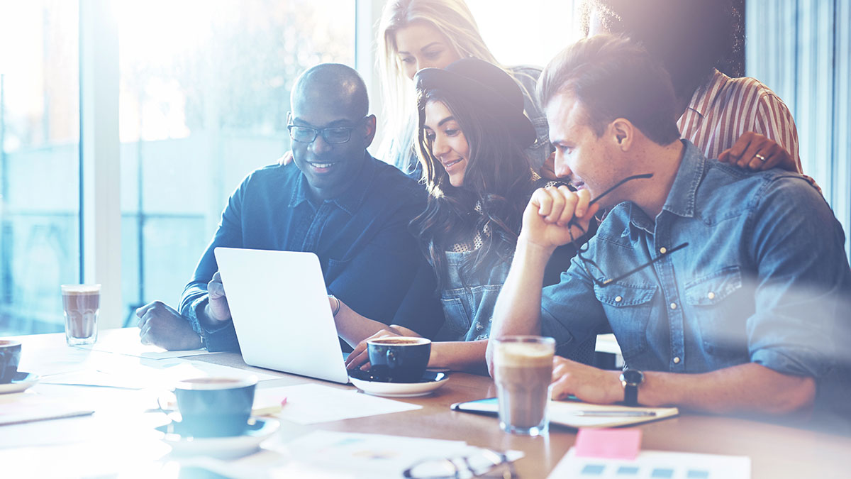 A diverse group of five people collaborates cheerfully around a laptop in a bright office, with coffee cups on the table, conveying teamwork and creativity.