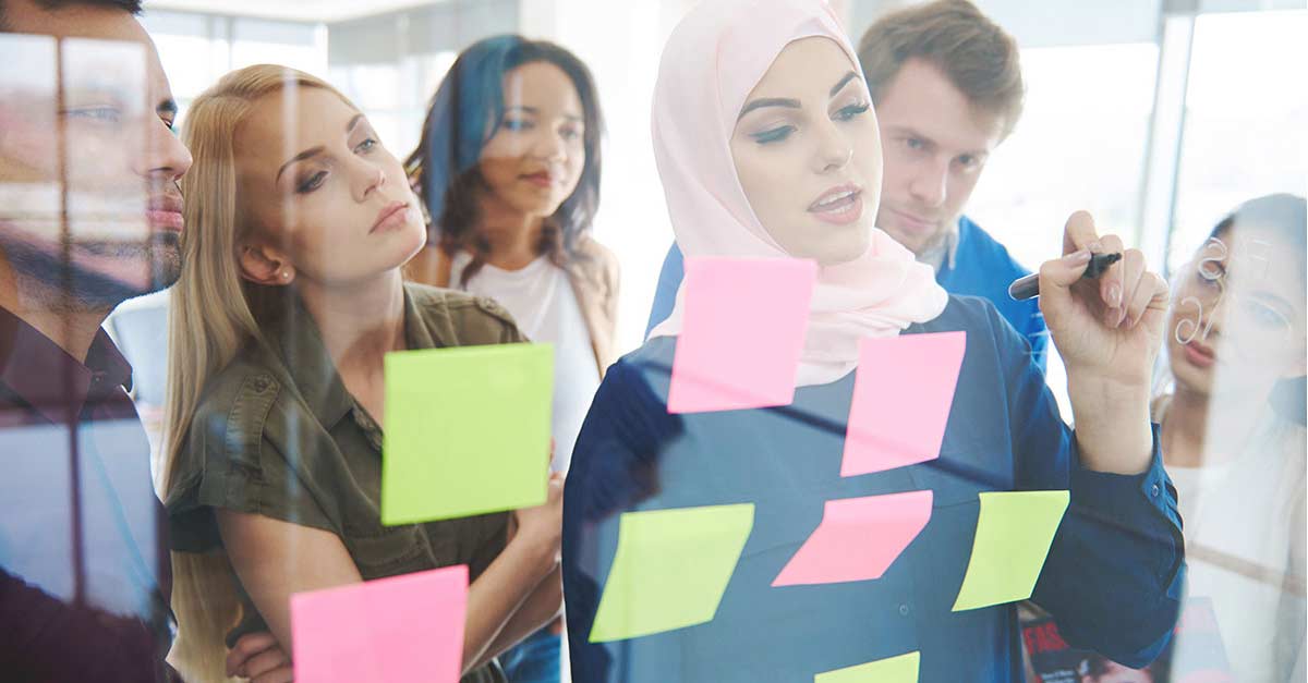 A diverse group of people in a modern office collaborate, focused on colorful sticky notes on a glass wall. The atmosphere is engaged and thoughtful.