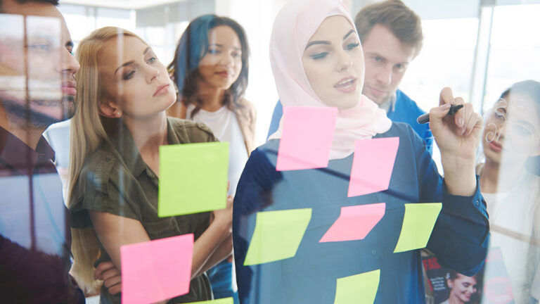 A diverse group of six people brainstorms around a glass wall covered in colorful sticky notes. A woman in a hijab writes with focus, creating a collaborative and innovative atmosphere.