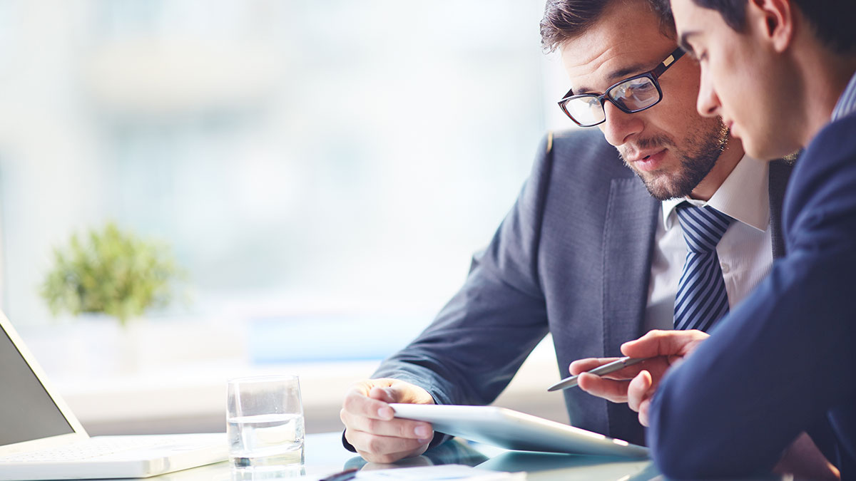 Two businessmen in suits, engaged in discussion over a tablet, sit at a bright office desk. A laptop, glass of water, and plant are visible nearby.