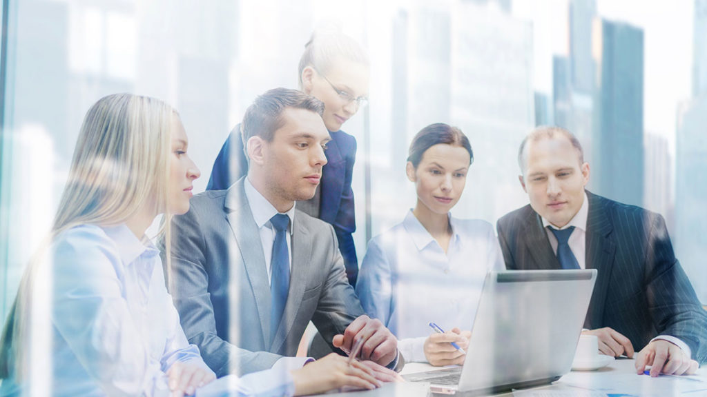 A diverse group of five professionals in business attire gather around a laptop in a modern office, focused and engaged in a collaborative discussion.