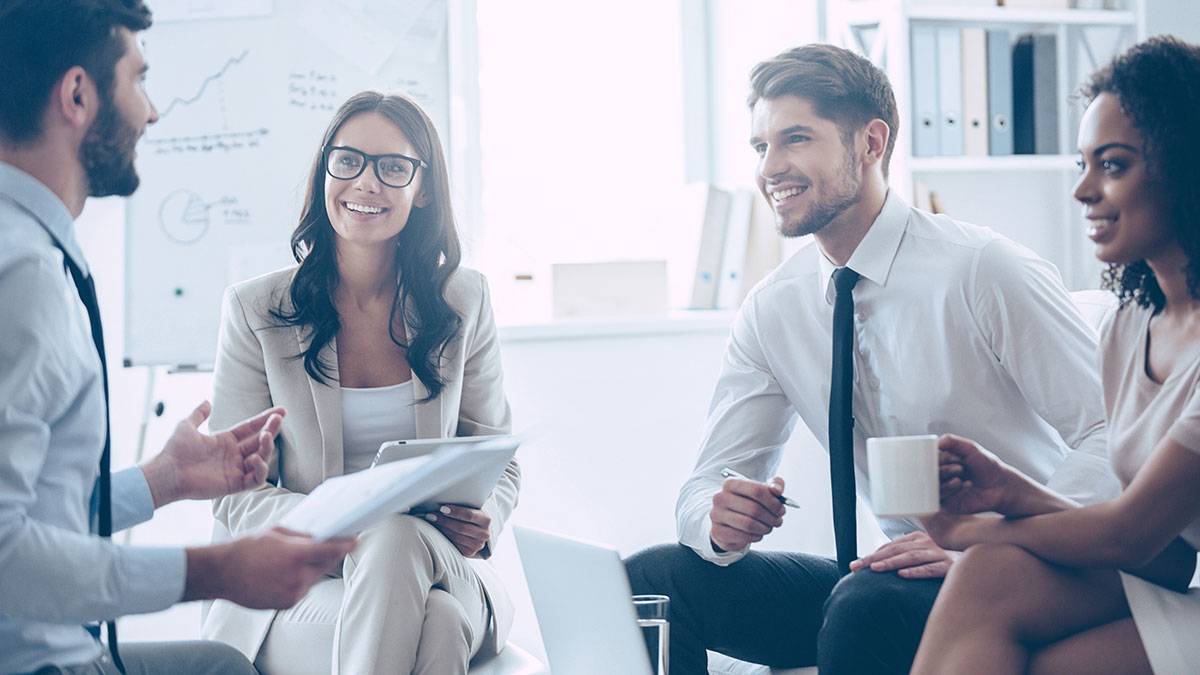 A diverse group of four business people in a bright office, smiling and engaged in discussion. One holds a tablet, another a coffee mug. Charts in the background.
