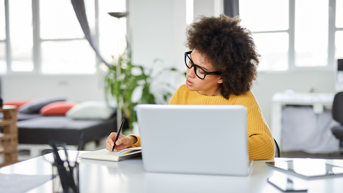 A woman in glasses and a yellow sweater writes in a notebook at a desk with a laptop. She's in a bright room with a modern, relaxed vibe.