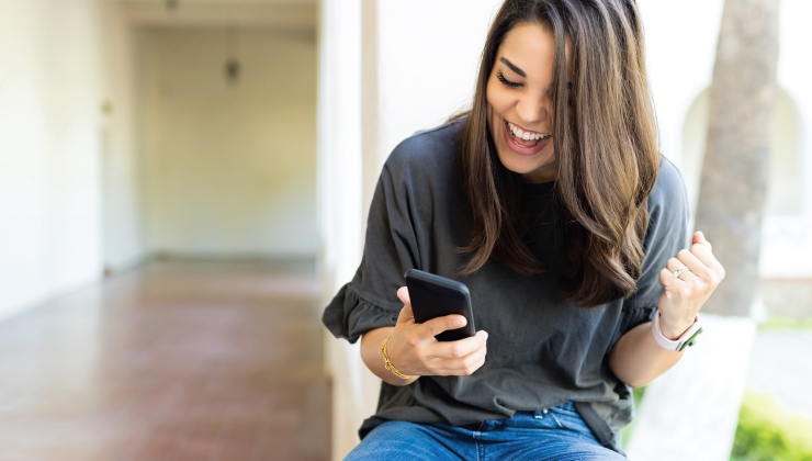 A woman sits and holds a smartphone, looking at it with excitement. She's outdoors, wearing a casual gray top and jeans, holding a fist in joy.