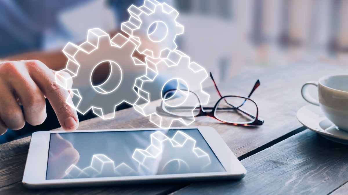 A person interacts with a tablet displaying digital gears, symbolizing technology and innovation. Nearby are eyeglasses and a cup, suggesting a work setting.