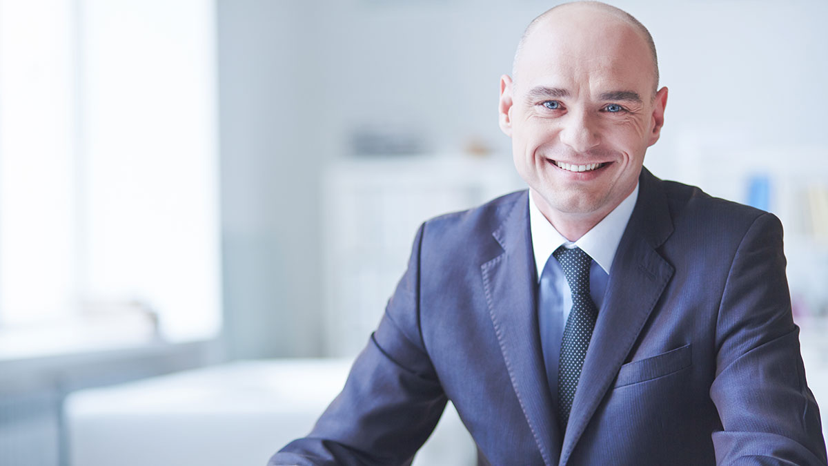 Smiling man in a suit sitting in a bright office. The background is softly blurred, creating a professional and welcoming atmosphere.